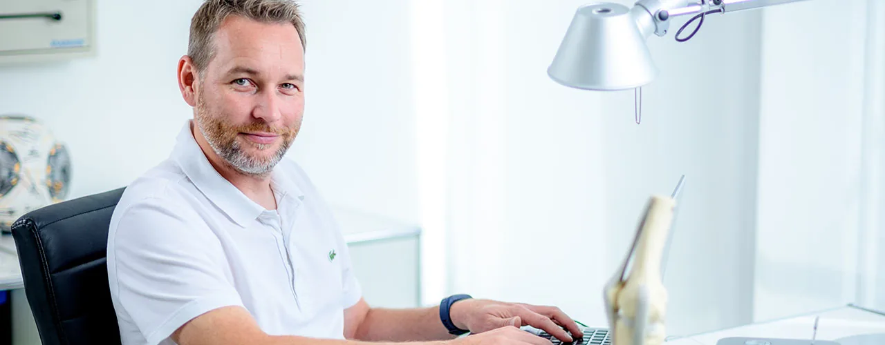 Arzt in weißem Poloshirt sitzt im hellen Sprechzimmer einer orthopädischen Praxis.